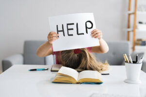 A young girl holding up a sign that reads HELP with an open school book. Get the help your child needs with learning disability testing in Berkeley, CA. Discover more here!
